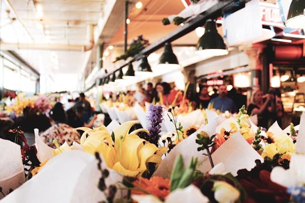 A bustling indoor market features an array of vibrant flowers wrapped in white paper. Shoppers walk through the aisles under industrial-style lighting. The environment is lively, filled with people exploring the fresh and colorful floral arrangements.