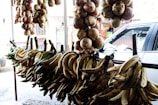 Bunches of plantains and onions are hanging in an outdoor market setup. A parked car is visible in the background, along with a few stalls.