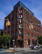 A multi-story brick building with large windows and a vertical sign displaying the word 'Hotel'. The structure has a modern, industrial design with metal and glass elements on the rooftop. The street in front features a cyclist riding by and a few pedestrians walking near parked cars. The setting includes traffic signals and a clear, sunny sky.