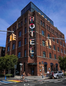 A multi-story brick building with large windows and a vertical sign displaying the word 'Hotel'. The structure has a modern, industrial design with metal and glass elements on the rooftop. The street in front features a cyclist riding by and a few pedestrians walking near parked cars. The setting includes traffic signals and a clear, sunny sky.