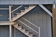 An outdoor staircase with wooden steps and railings set against a vertical paneling wall. The structure appears to be part of a building exterior, with muted tones and a simple design.