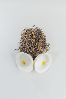 A cluster of dry tea leaves is spread on a surface, accompanied by two small white ceramic cups with a hint of tea inside.