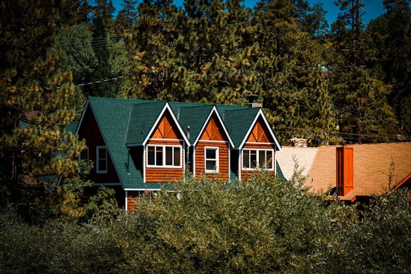 A charming wooden house is nestled among lush green pine trees. The house features a steep, dark green roof and multiple windows on the upper floor with white frames. Its warm wood paneling contrasts with the surrounding greenery. A sloped, light brown roof of a nearby structure is partially visible.
