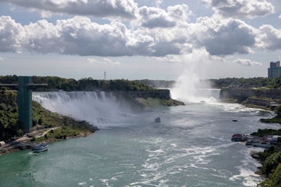 A majestic view of Niagara Falls with cascading water creating mist. The falls are flanked by lush greenery, with boats visible in the water below. A tower can be seen to the left, and there are clouds scattered across the sky.