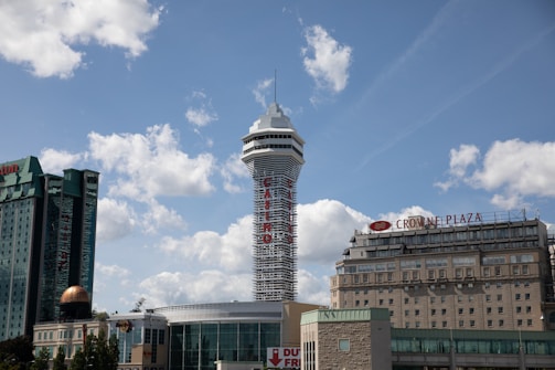 Several high-rise buildings are visible under a bright sky with fluffy clouds. The tallest structure has a unique tower design with a casino sign. Other buildings include a hotel with a distinctive green facade and another with a Crowne Plaza sign.