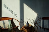 A cozy indoor café scene bathed in warm light, featuring two brown leather chairs with striped cushions. A shadow on the wall displays the word 'ESPRESSO' while a small table holds a bottle and a plant with bare branches.