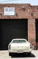 A vintage white car is parked in front of a large, closed, black industrial garage door. The building is made of red bricks, and a sign above the garage door reads 'FOR LEASE' with a phone number. The scene has an urban, slightly neglected look.