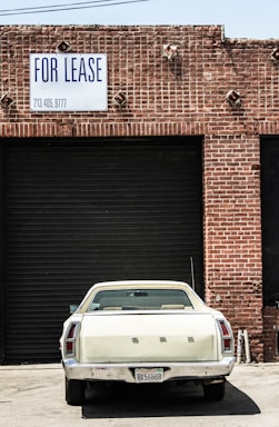 A vintage white car is parked in front of a large, closed, black industrial garage door. The building is made of red bricks, and a sign above the garage door reads 'FOR LEASE' with a phone number. The scene has an urban, slightly neglected look.