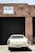 A vintage white car is parked in front of a large, closed, black industrial garage door. The building is made of red bricks, and a sign above the garage door reads 'FOR LEASE' with a phone number. The scene has an urban, slightly neglected look.