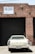 A vintage white car is parked in front of a large, closed, black industrial garage door. The building is made of red bricks, and a sign above the garage door reads 'FOR LEASE' with a phone number. The scene has an urban, slightly neglected look.