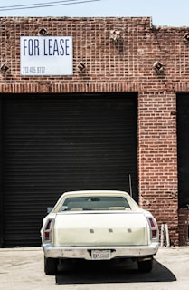 A vintage white car is parked in front of a large, closed, black industrial garage door. The building is made of red bricks, and a sign above the garage door reads 'FOR LEASE' with a phone number. The scene has an urban, slightly neglected look.