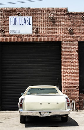 A vintage white car is parked in front of a large, closed, black industrial garage door. The building is made of red bricks, and a sign above the garage door reads 'FOR LEASE' with a phone number. The scene has an urban, slightly neglected look.