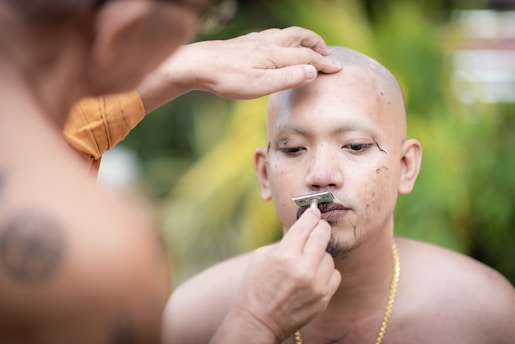A monk is shaving the head of a man outdoors. The man is seated with a focused expression as another person uses a razor to shave his facial hair. The scene is set against a blurred, natural green background, suggesting a serene and peaceful environment.