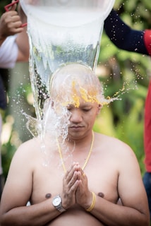 A shirtless person kneels with eyes closed and hands in a prayer position while water is being poured over their shaved head. The person wears a gold chain and a wristwatch. Two hands visible on either side are pouring water from a large container. The background appears to be lush and green, suggesting an outdoor setting.