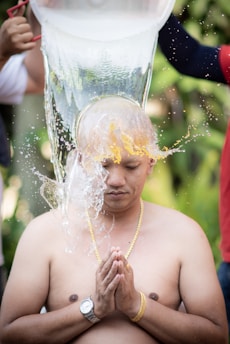 A shirtless person kneels with eyes closed and hands in a prayer position while water is being poured over their shaved head. The person wears a gold chain and a wristwatch. Two hands visible on either side are pouring water from a large container. The background appears to be lush and green, suggesting an outdoor setting.
