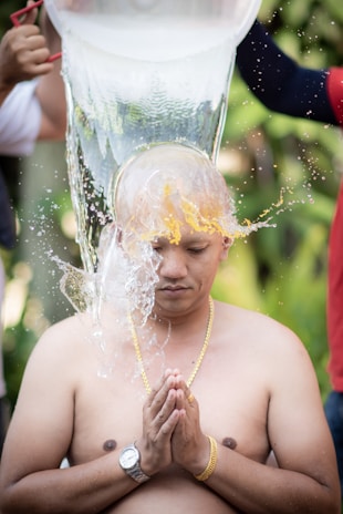 A shirtless person kneels with eyes closed and hands in a prayer position while water is being poured over their shaved head. The person wears a gold chain and a wristwatch. Two hands visible on either side are pouring water from a large container. The background appears to be lush and green, suggesting an outdoor setting.