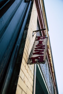 A wooden sign with the word 'Pizza' in red letters hangs from a chain on the side of a brick building. The building has a classic, slightly worn look with a combination of yellow brick and dark trim. The angle is upward, giving a dramatic view of the sign and the building's edge against a clear blue sky.