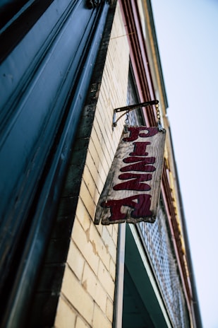 A wooden sign with the word 'Pizza' in red letters hangs from a chain on the side of a brick building. The building has a classic, slightly worn look with a combination of yellow brick and dark trim. The angle is upward, giving a dramatic view of the sign and the building's edge against a clear blue sky.
