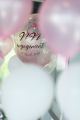 A transparent balloon with text and a pink ring graphic has 'engagement' and a date printed on it. It is surrounded by other balloons with a soft focus, primarily in pink and white hues.