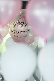 A transparent balloon with text and a pink ring graphic has 'engagement' and a date printed on it. It is surrounded by other balloons with a soft focus, primarily in pink and white hues.