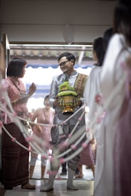 A man in traditional attire is holding a decorative container and is smiling at a woman also in traditional clothing. They are surrounded by a festive atmosphere with decorative ropes adorned with pink and white flowers. Several people in the background appear to be part of a celebratory gathering.