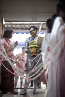 A man in traditional attire is holding a decorative container and is smiling at a woman also in traditional clothing. They are surrounded by a festive atmosphere with decorative ropes adorned with pink and white flowers. Several people in the background appear to be part of a celebratory gathering.