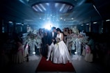 A wedding scene captures a bride and groom walking down a red carpet, surrounded by a dimly lit venue. The bride is wearing a white wedding dress, holding a bouquet of flowers, while the groom is dressed in a dark suit and white pants. Guests stand around them, some in elegant dresses. Large floral arrangements with white and pastel flowers adorn the sides.