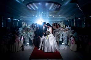 A wedding scene captures a bride and groom walking down a red carpet, surrounded by a dimly lit venue. The bride is wearing a white wedding dress, holding a bouquet of flowers, while the groom is dressed in a dark suit and white pants. Guests stand around them, some in elegant dresses. Large floral arrangements with white and pastel flowers adorn the sides.