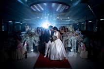 A wedding scene captures a bride and groom walking down a red carpet, surrounded by a dimly lit venue. The bride is wearing a white wedding dress, holding a bouquet of flowers, while the groom is dressed in a dark suit and white pants. Guests stand around them, some in elegant dresses. Large floral arrangements with white and pastel flowers adorn the sides.