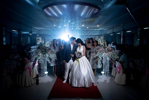 A wedding scene captures a bride and groom walking down a red carpet, surrounded by a dimly lit venue. The bride is wearing a white wedding dress, holding a bouquet of flowers, while the groom is dressed in a dark suit and white pants. Guests stand around them, some in elegant dresses. Large floral arrangements with white and pastel flowers adorn the sides.