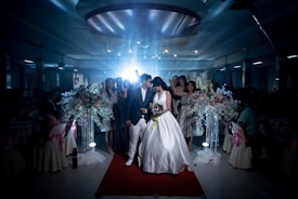 A wedding scene captures a bride and groom walking down a red carpet, surrounded by a dimly lit venue. The bride is wearing a white wedding dress, holding a bouquet of flowers, while the groom is dressed in a dark suit and white pants. Guests stand around them, some in elegant dresses. Large floral arrangements with white and pastel flowers adorn the sides.