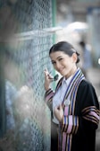 A person in graduation attire poses beside a chain-link fence. The outfit features colorful stripes and an emblem, and the individual is holding sunglasses and smiling warmly.