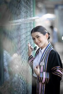A person in graduation attire poses beside a chain-link fence. The outfit features colorful stripes and an emblem, and the individual is holding sunglasses and smiling warmly.