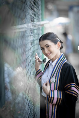 A person in graduation attire poses beside a chain-link fence. The outfit features colorful stripes and an emblem, and the individual is holding sunglasses and smiling warmly.