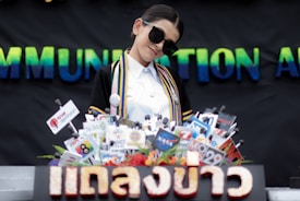 A person dressed in an academic gown and sunglasses is standing behind a podium covered with numerous microphones featuring various logos and labels. The background displays the word 'COMMUNICATION' in colorful letters.