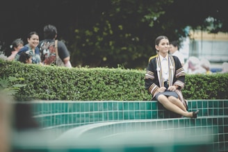 A person in graduation attire sits on the edge of a tiled water feature with vibrant greenery in the background. Other people are present, some smiling and interacting in a relaxed outdoor setting.