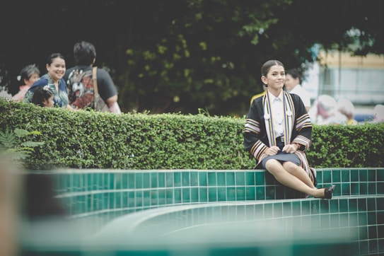 A person in graduation attire sits on the edge of a tiled water feature with vibrant greenery in the background. Other people are present, some smiling and interacting in a relaxed outdoor setting.