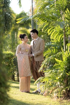 A couple stands together in a lush garden setting, with both wearing traditional attire. They are gazing at each other warmly, surrounded by vibrant green foliage and bathed in soft, natural light.