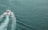A motorboat travels across a vast expanse of clear, greenish-blue ocean water, leaving a foamy wake behind it as it propels forward. The boat features a cabin and is powered by two outboard motors. The water's surface is slightly textured with gentle waves.