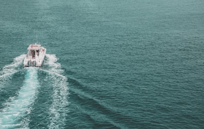 A motorboat travels across a vast expanse of clear, greenish-blue ocean water, leaving a foamy wake behind it as it propels forward. The boat features a cabin and is powered by two outboard motors. The water's surface is slightly textured with gentle waves.