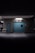A dimly lit industrial interior features a large blue sliding door flanked by metal siding. Above the door, there is a bright white light fixture. To the left, a small blue service door is present. A bicycle is visible in the shadowy area to the far left, suggesting a parking or storage space.