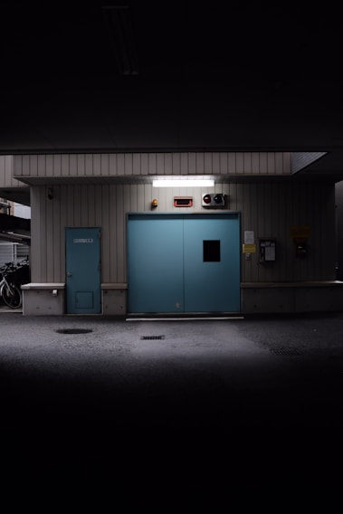 A dimly lit industrial interior features a large blue sliding door flanked by metal siding. Above the door, there is a bright white light fixture. To the left, a small blue service door is present. A bicycle is visible in the shadowy area to the far left, suggesting a parking or storage space.