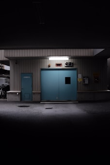 A dimly lit industrial interior features a large blue sliding door flanked by metal siding. Above the door, there is a bright white light fixture. To the left, a small blue service door is present. A bicycle is visible in the shadowy area to the far left, suggesting a parking or storage space.