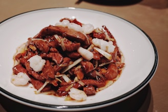 A plate of stir-fried meat and vegetables, with pieces of white seafood or tofu garnished among the slices. The dish appears to be seasoned with a dark sauce that coats the ingredients. The meal is served on a white plate with a dark rim, placed on a brown surface.