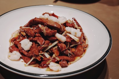 A plate of stir-fried meat and vegetables, with pieces of white seafood or tofu garnished among the slices. The dish appears to be seasoned with a dark sauce that coats the ingredients. The meal is served on a white plate with a dark rim, placed on a brown surface.