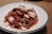 A plate of stir-fried meat and vegetables, with pieces of white seafood or tofu garnished among the slices. The dish appears to be seasoned with a dark sauce that coats the ingredients. The meal is served on a white plate with a dark rim, placed on a brown surface.