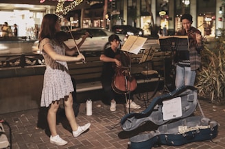 A group of three musicians perform on a street corner in an urban setting at night. Two individuals are playing violins while the third sits on a bench playing a cello. Music stands are positioned in front of the musicians, and a blue instrument case lies open on the ground, containing money and a sign reading 'Support our fund'. Decorative lights and bustling pedestrians in a shopping area can be seen in the background.