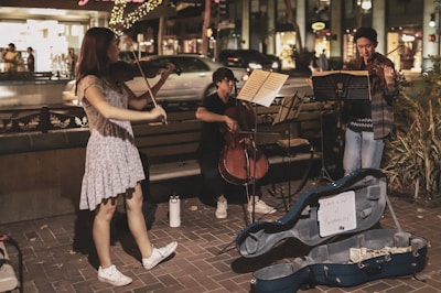 A group of three musicians perform on a street corner in an urban setting at night. Two individuals are playing violins while the third sits on a bench playing a cello. Music stands are positioned in front of the musicians, and a blue instrument case lies open on the ground, containing money and a sign reading 'Support our fund'. Decorative lights and bustling pedestrians in a shopping area can be seen in the background.
