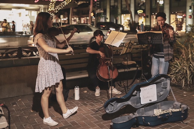 A group of three musicians perform on a street corner in an urban setting at night. Two individuals are playing violins while the third sits on a bench playing a cello. Music stands are positioned in front of the musicians, and a blue instrument case lies open on the ground, containing money and a sign reading 'Support our fund'. Decorative lights and bustling pedestrians in a shopping area can be seen in the background.
