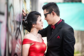 A couple stands close together in an intimate pose. The woman is wearing a red off-shoulder dress with elegant jewelry, her hair styled neatly. The man is dressed in a black suit with a red bow tie and pocket square, wearing sunglasses. They are positioned next to a graffiti-covered wall, with the man leaning against it, creating a romantic and stylish scene.
